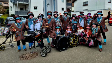 Eine bunte Gruppe von Karnevalisten in kostümierten Outfits steht zusammen. Sie tragen Hüte und haben ihre Gesichter geschminkt, während einige Musikinstrumente zu sehen sind.