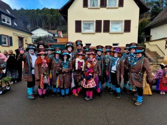 Eine Gruppe von fröhlichen Menschen in bunten Kostümen steht auf einer Straße vor einem Gebäude. Sie tragen Hüte und viele haben bemalte Gesichter.