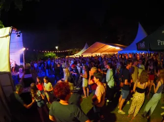 Eine lebhafte Veranstaltung bei Nacht mit einer großen Menschenmenge, die tanzt und feiert. Bunte Lichter und Zelte schaffen eine festliche Atmosphäre.