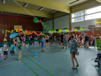 Ein bunt geschmückter Raum mit vielen Menschen, die feiern. Kinder spielen mit Ballons und es gibt eine fröhliche Atmosphäre.