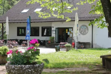 Ein gemütliches Café mit einer einladenden Terrasse und schönen Blumen. Im Hintergrund sind Bäume und eine ruhige Grünfläche zu sehen.