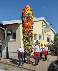 Eine Gruppe von Menschen steht um einen bunten, geschmückten Baum. Einige tragen traditionelle Kleidung und scheinen eine Feier zu veranstalten.
