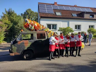 Eine Gruppe von Männern in roten Schürzen steht neben einem dekorierten, camouflagefarbenen Auto. Im Hintergrund ist ein Wohnhaus mit Solarpanelen zu sehen.