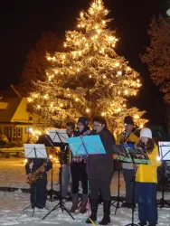 Eine Gruppe von Musikern spielt Instruments vor einem festlich beleuchteten Weihnachtsbaum. Der Schnee auf dem Boden ergänzt die winterliche Atmosphär.