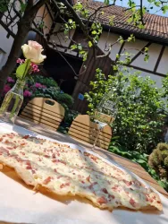 Eine frisch gebackene Flammkuchen liegt auf einem Tisch im Freien. Im Hintergrund sieht man eine grüne Umgebung mit Blumen und einem Glas Wein.