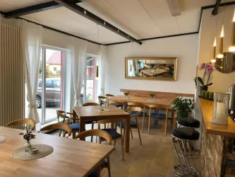Ein modernes Café mit hellen Wänden und Holzdekor. Es gibt mehrere Tische und Stühle sowie große Fenster, die viel Tageslicht hereinlassen.