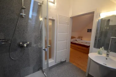 Ein modernes Badezimmer mit einer begehbaren Dusche und einem Waschbecken. Im Hintergrund ist ein Schlafzimmer mit einem Bett sichtbar.