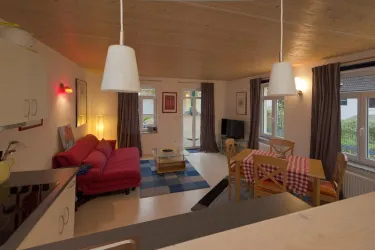 Ein gemütlicher Wohnbereich mit einem roten Sofa und einem Esstisch. Große Fenster sorgen für viel Tageslicht.