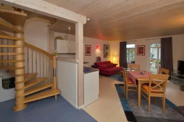 Eine helle und moderne Wohnung mit einer Holztreppe und einem gemütlichen Wohnbereich. Es gibt einen Esstisch und einen roten Sofa, sowie große Fenster, die viel Tageslicht hereinlassen.