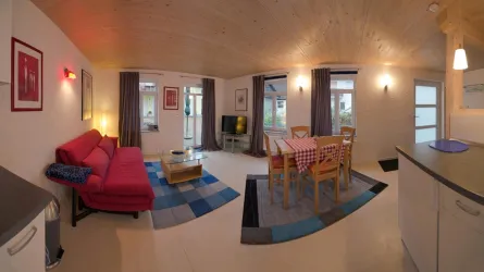 Ein modernes Wohnzimmer mit einem roten Sofa und einem Essbereich. Große Fenster sorgen für viel Tageslicht und eine gemütliche Atmosphäre.