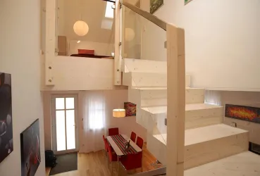 Eine moderne Innenansicht eines Hauses mit Holztreppe. Im Vordergrund befindet sich ein Essbereich mit roten Stühlen und einem langen Tisch.