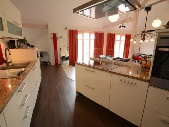 Eine moderne Küche mit hellen Oberflächen und Granitplatten. Große Fenster lassen viel Tageslicht herein und bieten einen Blick auf den Wohnbereich.