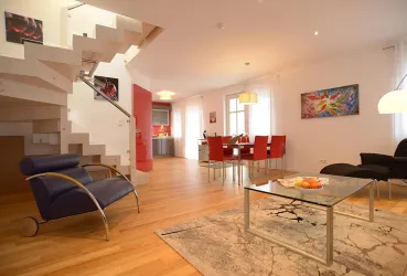 Ein modernes Wohnzimmer mit einem klaren, offenen Raum. Die Einrichtung umfasst einen blauen Sessel, einen Glastisch und einen Esstisch mit roten Stühlen.