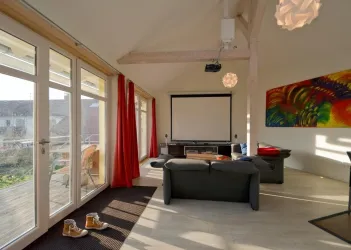 Ein modernes Wohnzimmer mit großen Fenstern und rotem Vorhang. Im Hintergrund steht ein Fernseher und es gibt bunte Kunst an der Wand.