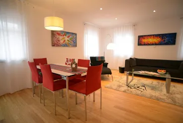 Ein heller Wohnraum mit einem Essbereich und roten Stühlen. Im Hintergrund stehen ein schwarzes Sofa und kunstvolle Wandbilder.
