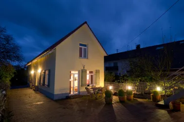Ein schönes, gelbes Haus bei Nacht mit warmem Licht. Im Vordergrund sind Tische und Pflanzen zu sehen.
