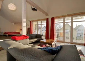 Ein modernes Wohnzimmer mit großen Fenstern und viel natürlichem Licht. Es gibt gemütliche Sofas und einen Tisch, sowie rote Vorhänge, die einen warmen Akzent setzen.
