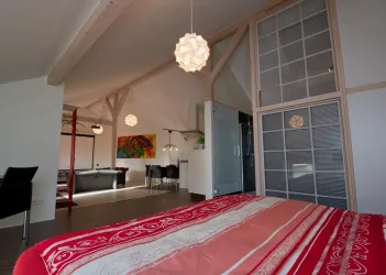 Ein modernes Schlafzimmer mit einem roten Bett und einer hellen Decke. Im Hintergrund sind ein gemütlicher Wohnbereich und kunstvolle Leuchten sichtbar.