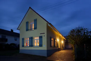 Ein gelbes Haus mit blauen Fensterläden steht bei Dämmerung. Die Fenster sind beleuchtet und schaffen eine gemütliche Atmosphäre.
