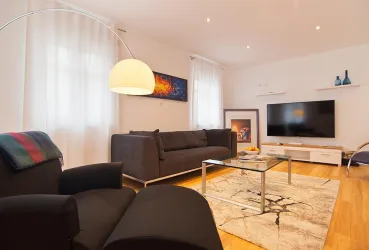 Ein modernes Wohnzimmer mit einem schwarzen Sofa und einem Sessel. Der Raum ist hell und hat einen Fernseher an der Wand.