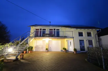 Ein modernes Haus mit einer hellen Fassade und einem gepflasterten Vorplatz. Es ist Nacht und die Fenster sind beleuchtet.