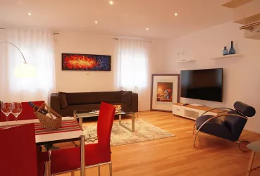 Ein modernes Wohnzimmer mit einem Sofa, einem Fernseher und einem Esstisch. Die Wände sind hell gestrichen, und es gibt dekorative Kunstwerke sowie einen warmen Holzboden.