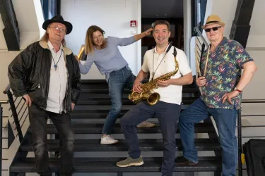 Eine Gruppe von vier Musikern steht auf einer Treppe. Sie posieren fröhlich mit verschiedenen Musikinstrumenten.