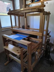 Ein Webstuhl aus Holz steht in einem hellen Raum. Auf dem Webstuhl liegt ein blauer Stoff, umgeben von Spulen mit Garn.