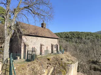 Eine alte Kapelle auf einem Hügel, umgeben von Bäumen. Der Himmel ist klar und blau, und die Landschaft ist grün.