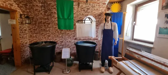 Ein rustikaler Raum mit rotem Backsteinmauerwerk, zwei großen Kesseln und einer Puppe in historischer Kleidung, ergänzt durch bunte Fahnen im Hintergrund.