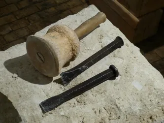 Ein Steinblock mit einem Holzhammer und zwei Meißeln aus Metall liegt darauf. Die Werkzeuge sind für handwerkliche Tätigkeiten gedacht, wie zum Beispiel für den Steinmetz.