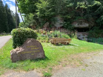 Ein malerischer Landschaftsbereich mit einem Hinweisstein und einer Sitzbank. Umgeben von grüner Vegetation und einem kleinen Felsvorsprung. Hier der Picknickplatz "Speyerbachquelle" von Speyerbrunn zu sehen.
