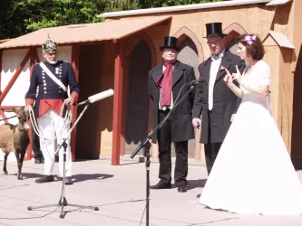 Eine festliche Veranstaltung mit vier Personen in formeller Kleidung und einer Ziege auf der Bühne. Der Hintergrund besteht aus einer dekorativen Kulisse.