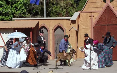 Eine Theateraufführung mit Menschen in historischen Kostümen. In der Mitte der Bühne steht eine Ziege, während die Darsteller interagieren.