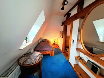 Ein gemütliches Zimmer mit einem Einzelbett und blauen Teppichboden. Es hat schräg verlaufende Fenster und Holzregale für eine warme Atmosphäre.