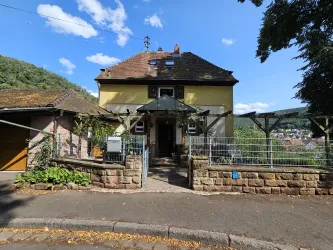 Ein charmantes Haus mit gelber Fassade und einem Vordach. Der Garten ist gepflegt und die Umgebung ist grün und hügelig.