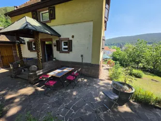 Eine Terrasse mit einem Tisch und Stühlen in einem ruhigen Garten. Im Hintergrund sind Bäume und Hügel zu sehen.