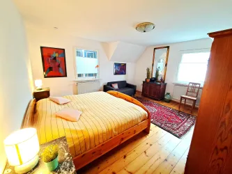 Ein gemütliches Schlafzimmer mit einem großen Bett und einem Teppich. Die Wände sind mit Kunstwerken dekoriert und es gibt einen kleinen Sitzbereich.