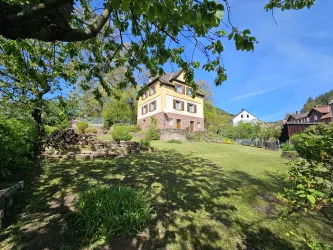 Ein großes, gelbes Haus steht auf einem Hügel umgeben von grünem Garten. Im Hintergrund sind weitere Häuser und blauer Himmel sichtbar.