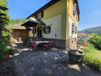Ein charmantes Haus mit gelber Fassade und einem überdachten Eingangsbereich. Draußen befindet sich eine Terrasse mit einem Tisch und Stühlen, umgeben von Grün.