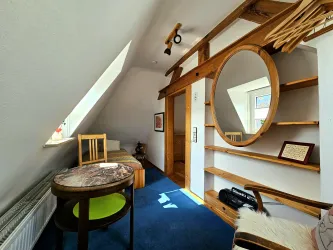 Ein gemütlicher Raum mit schrägen Wänden und einer großen runden Wandspiegel. Ein Tisch und ein Stuhl sowie offene Regale sind sichtbar.