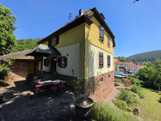 Ein charmantes Haus mit gelber Fassade und sichtbarem Garten. Im Hintergrund sind Bäume und Hügel zu sehen.