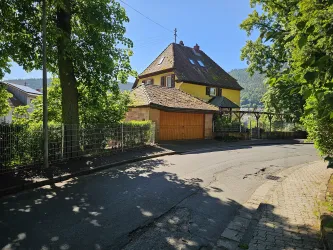 Ein gelbes Haus mit einem schrägen Dach steht an einer ruhigen Straße, umgeben von Bäumen. Die Sonne scheint und der Himmel ist klar.