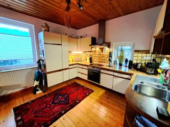 Eine moderne Küche mit Holzdecke und hellen Schränken. Ein Teppich liegt auf dem Holzboden, und an der Wand sind Küchenutensilien und Geräte zu sehen.