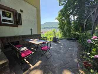 Eine einladende Terrasse mit einem Tisch und Stühlen, umgeben von grünen Bäumen. Im Hintergrund ist eine malerische Landschaft zu sehen.