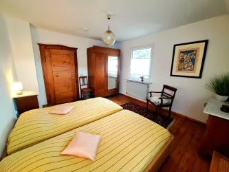 Ein gemütliches Schlafzimmer mit zwei Einzelbett und gelben Bettbezügen. Es gibt Holzmöbel, einen Stuhl und ein Fenster, das Tageslicht hereinlässt.