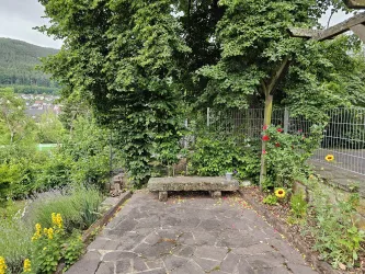 Ein idyllischer Garten mit einer Steinbank, umgeben von üppigem Grün. Bunte Blumen und ein Blick auf die sanften Hügel machen die Szenerie einladend.