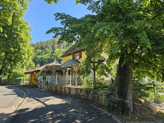 Ein schönes gelbes Haus, umgeben von Bäumen und grüner Landschaft. Die Straße führt sanft am Haus vorbei.