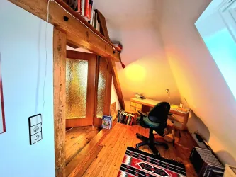 Ein gemütliches Arbeitszimmer mit Holzbalken und einem Schreibtisch. Im Raum befinden sich Bücherregale und ein Stuhl.