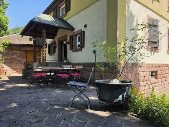 Eine gemütliche Terrasse mit einem Tisch und Stühlen in einem schönen alten Haus. Umgeben von Pflanzen und einem sonnigen Himmel.